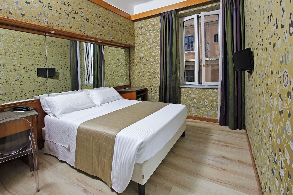 Habitación de hotel con cama, escritorio, teléfono, televisor y ventana con cortinas.