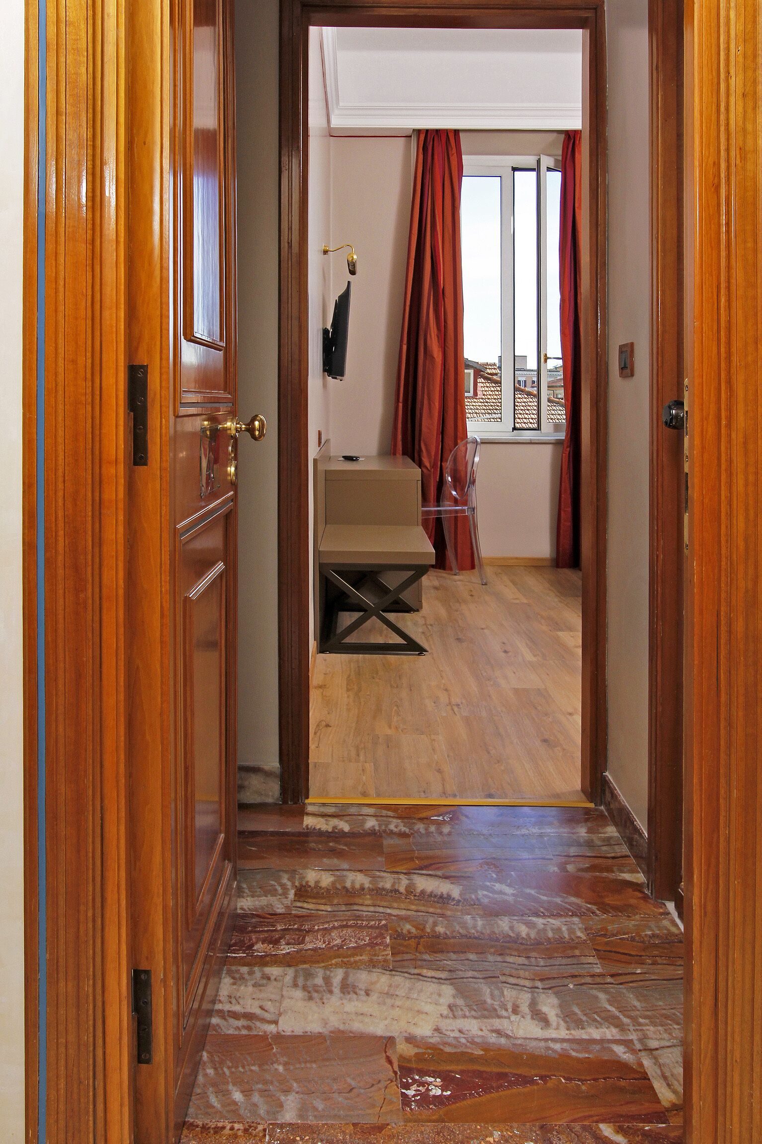 A wooden door leading to a room with a bed, a window with red curtains, and a view of buildings outside.
