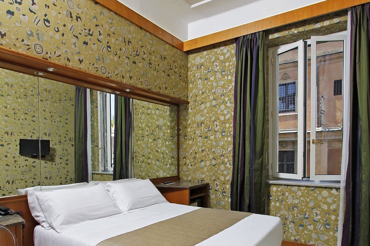 Habitación de hotel con cama, cabecero de madera, una mesita, una silla, televisor y ventana con cortinas.