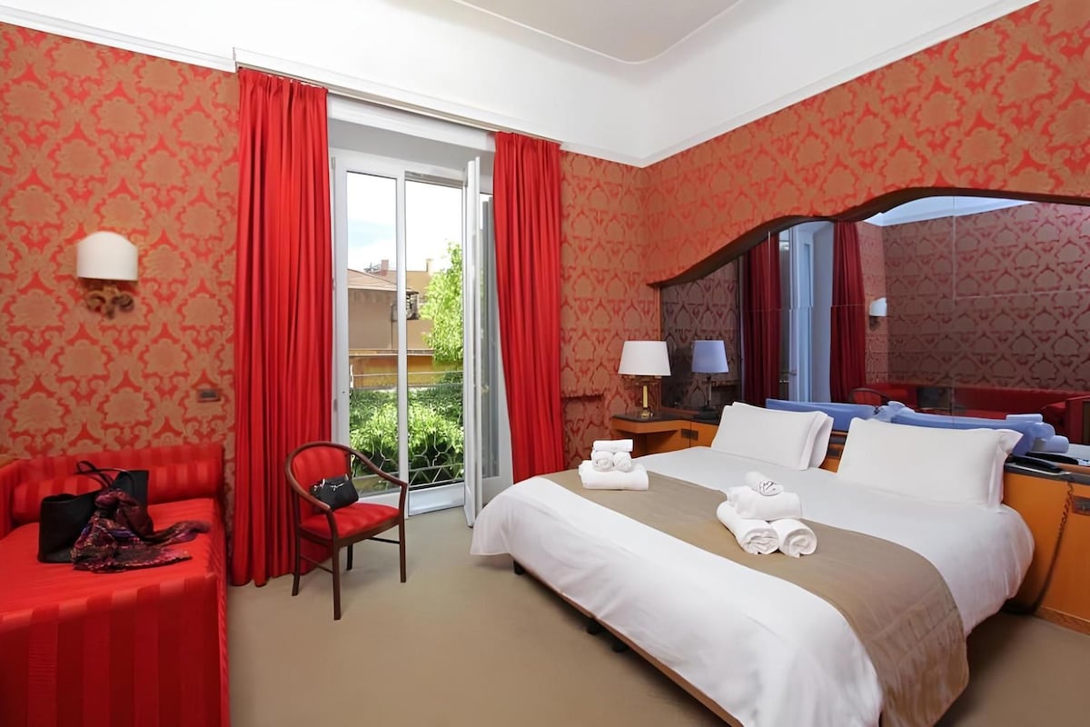 Een hotelkamer met een bed, een stoel, een bureau en een balkon met rode gordijnen.