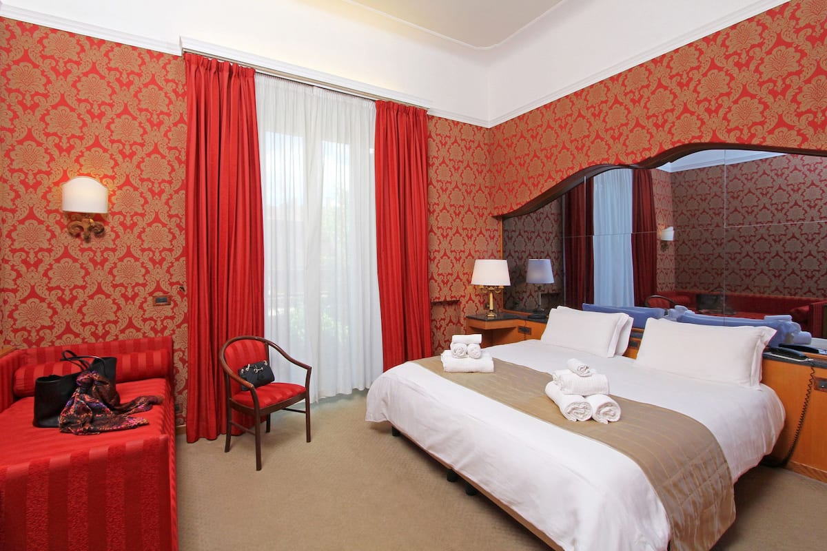 Habitación de hotel con una cama grande, un escritorio con silla, una lámpara de escritorio, un espejo y una ventana con cortinas rojas.