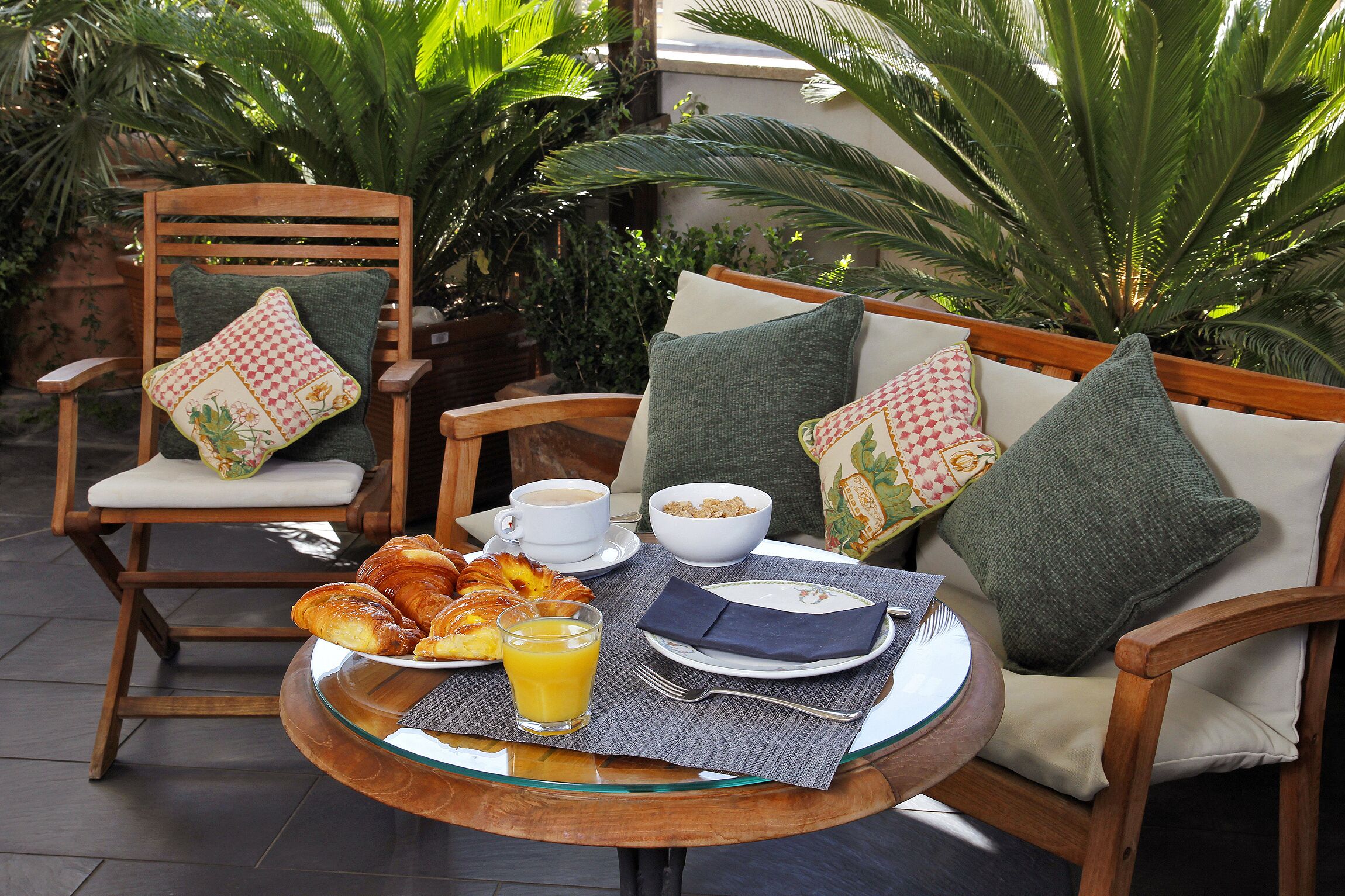 Une table en verre dressée pour le petit-déjeuner, avec des croissants, du jus d’orange et du café, entourée de chaises en bois dotées de coussins.