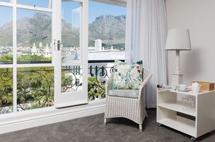 A balcony with a wicker chair, a white side table with a lamp, and a view of a cityscape and mountains.