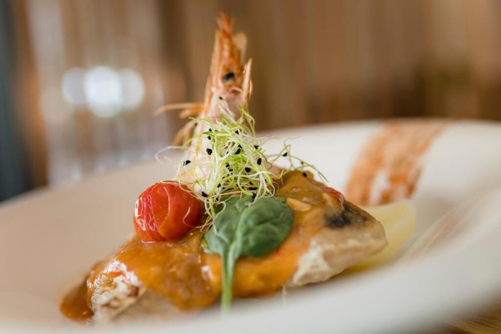 A plated dish with a piece of fish, a cherry tomato, a sprig of green, and a shrimp garnish.