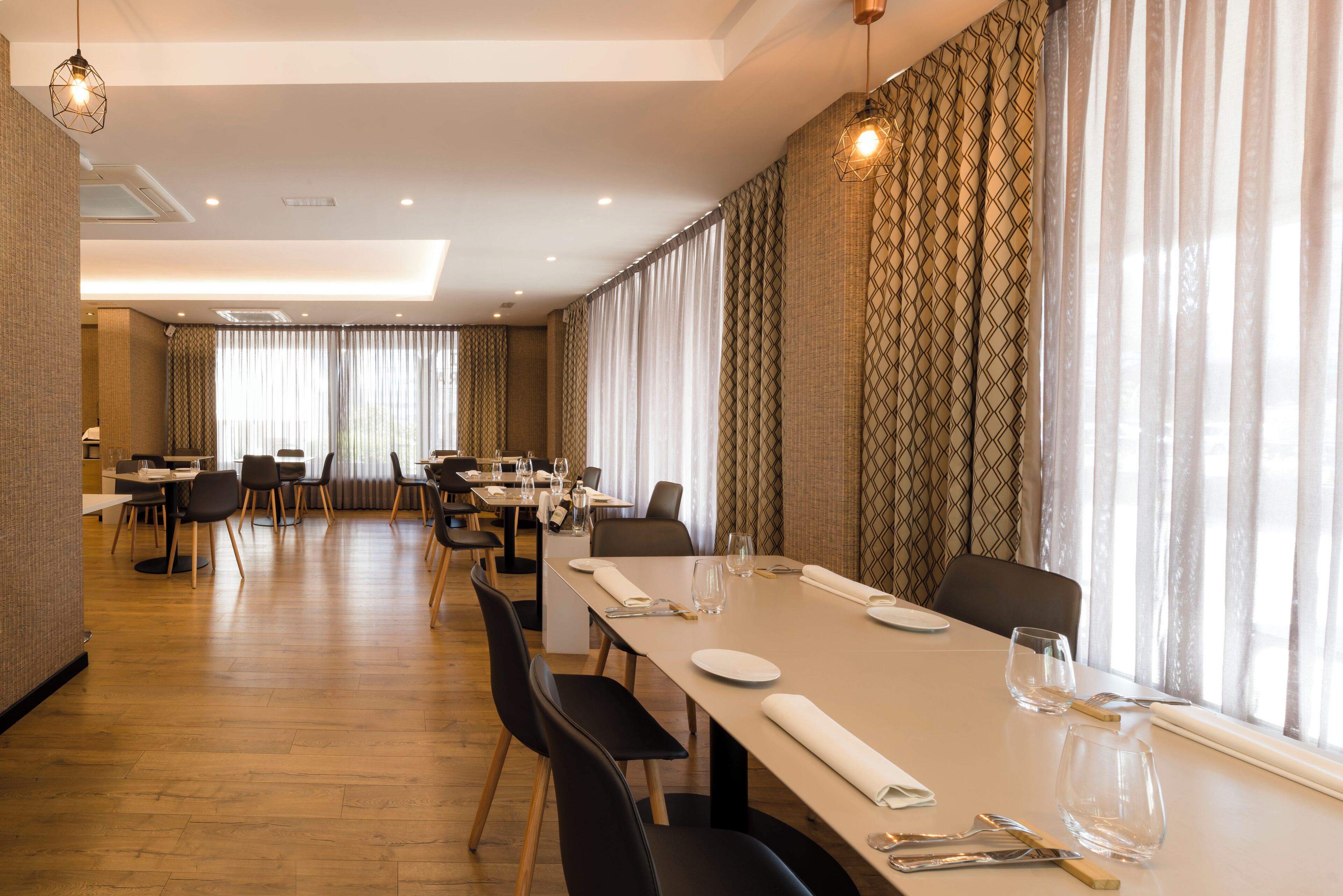 Un comedor moderno con mesas dispuestas para cuatro, suelo de madera y cortinas estampadas.