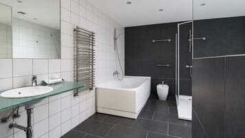 A modern bathroom with a glass sink, a white bathtub, a shower, and dark tiled walls.