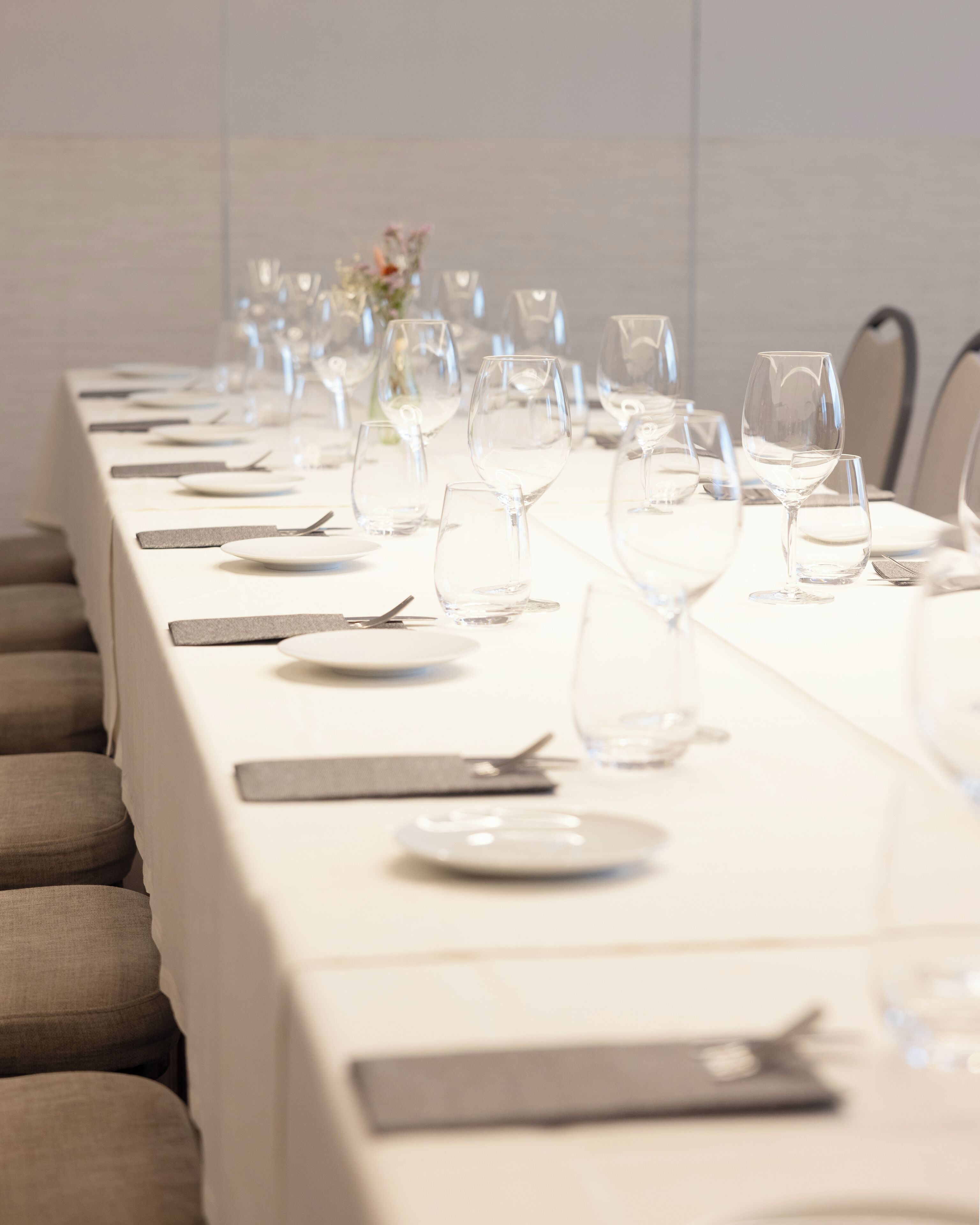 Una larga mesa de comedor dispuesta con varios vasos de vino, platos y cubiertos, lo que sugiere una formalidad en el servicio.