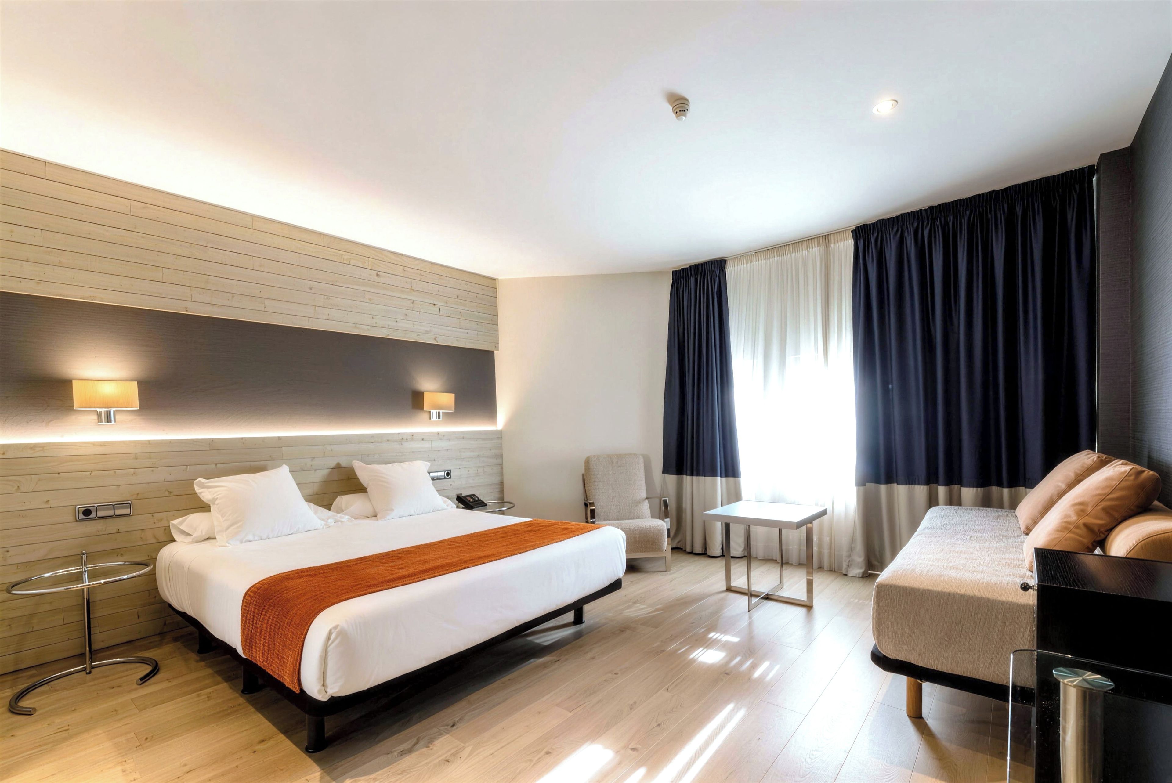 Una habitación de hotel moderna con una cama grande, mesitas de noche, un escritorio, una silla y un sofá.