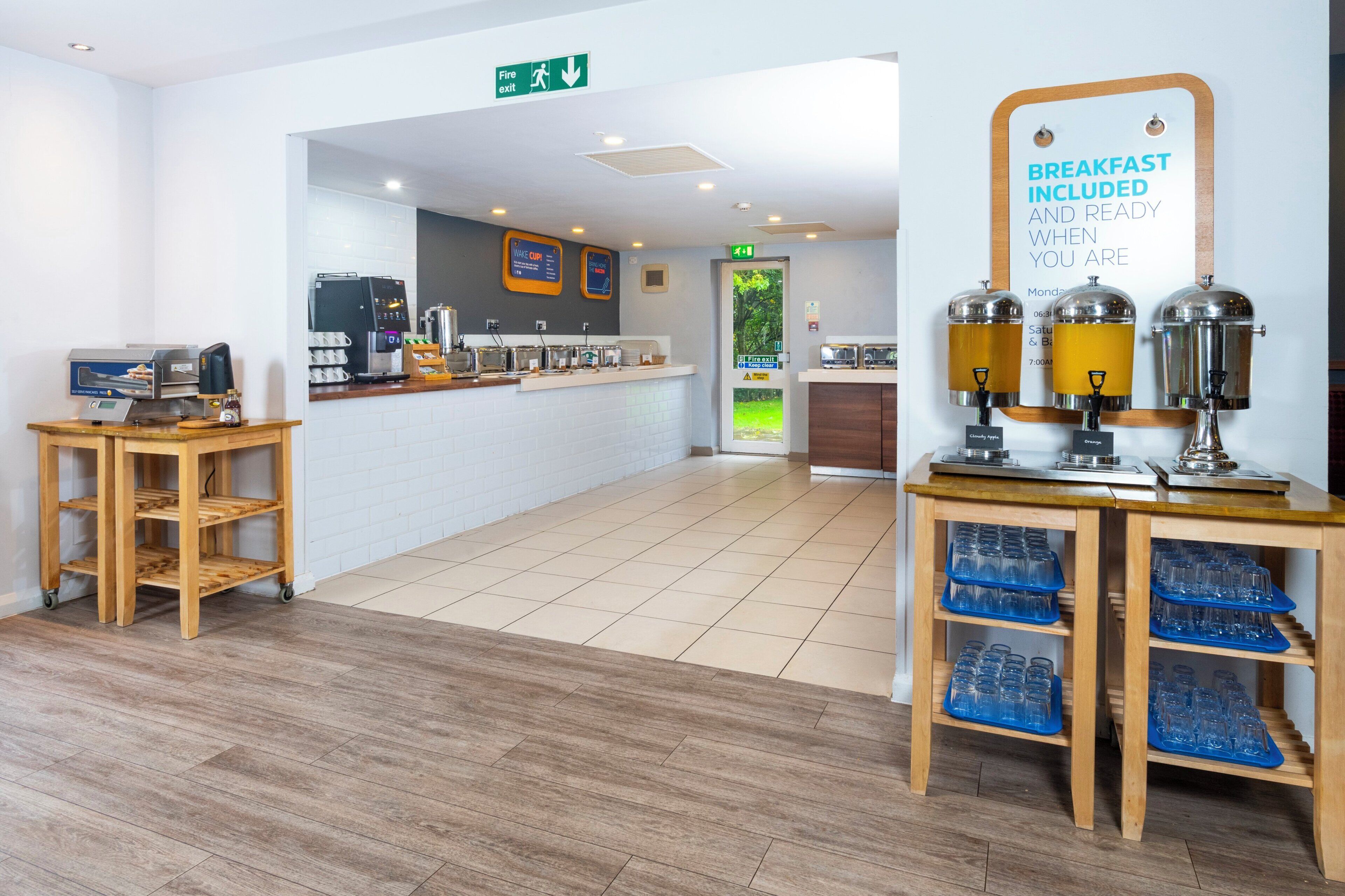 Un espace petit-déjeuner propre et moderne, équipé d’une machine à café, d’un panneau indiquant que le petit-déjeuner est inclus, et d’un panneau de signalisation pour la sortie de secours en cas d’incendie.