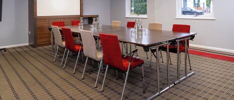 A conference room with a long table, red and beige chairs, a projector screen, and windows with a view of the outside.