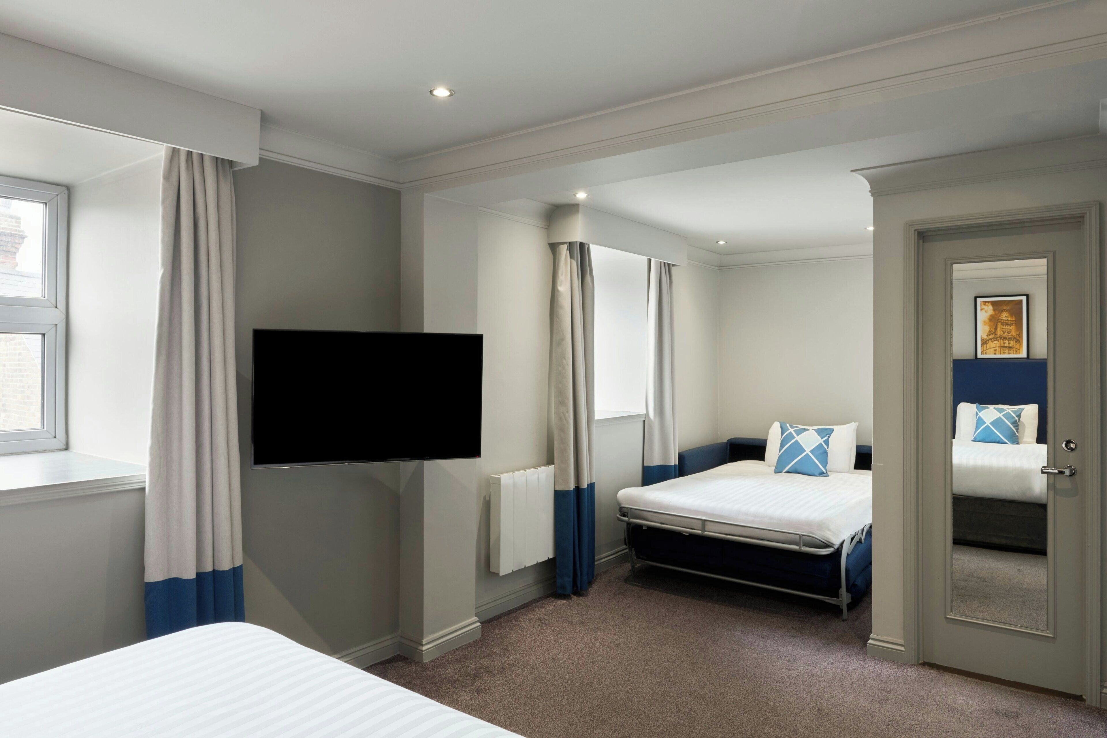 Ein Hotelzimmer mit einem Bett, einem an der Wand befestigten Fernseher, einem Fenster mit Vorhängen und einer Tür zu einem weiteren Zimmer.