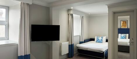 Ein Hotelzimmer mit einem Bett, einem an der Wand befestigten Fernseher, einem Fenster mit Vorhängen und einer Tür zu einem weiteren Zimmer.