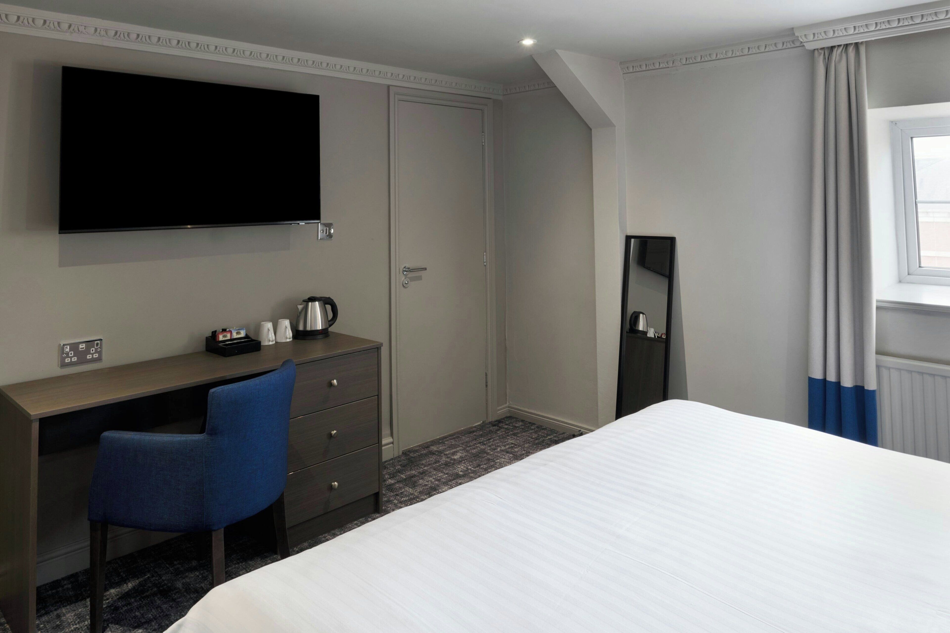 A hotel room with a bed, a desk with a chair, a television mounted on the wall, and a window with curtains.