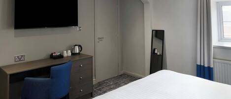 A hotel room with a bed, a desk with a chair, a television mounted on the wall, and a window with curtains.