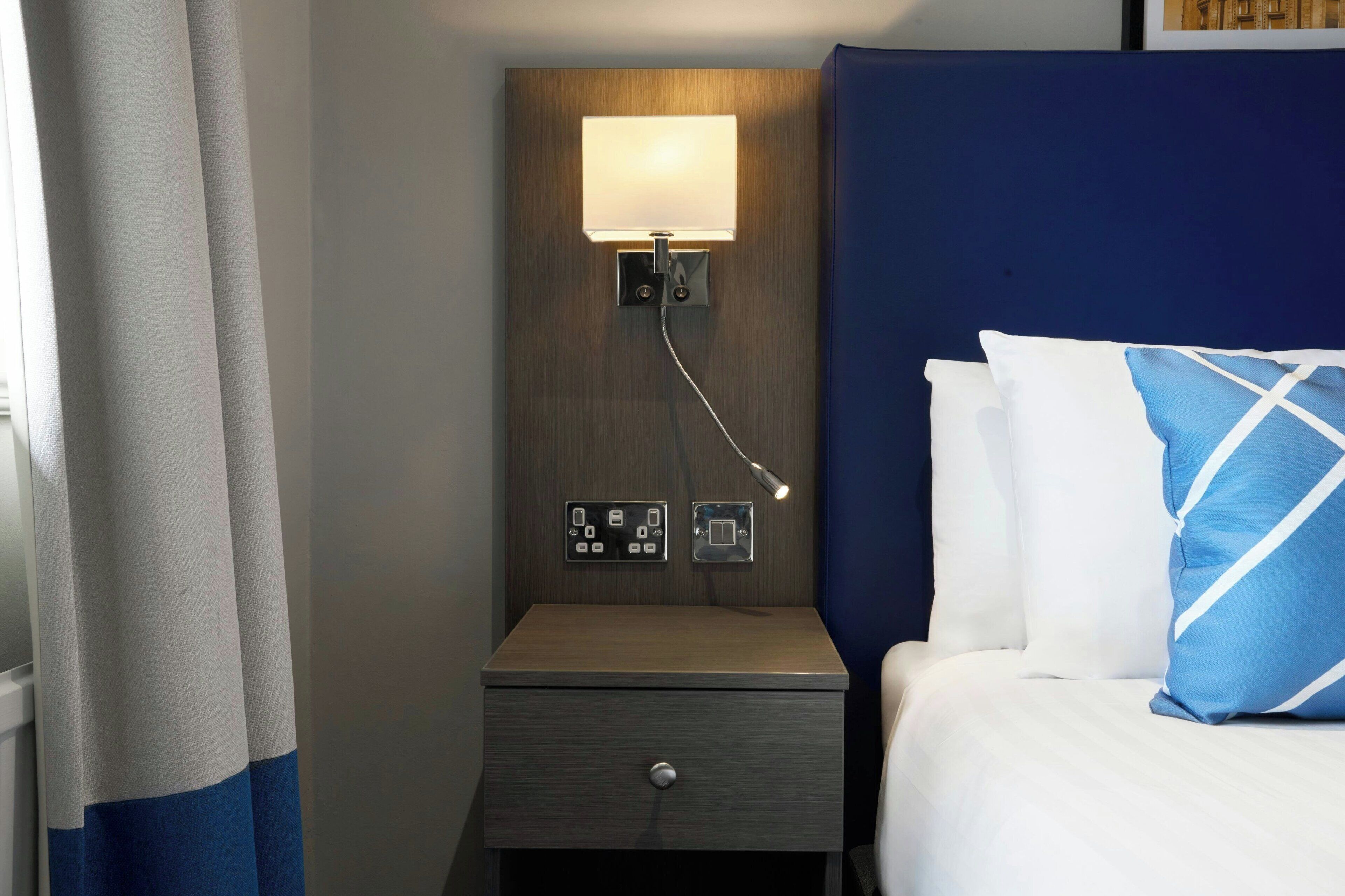 A hotel room with a bed, a nightstand with a lamp, and a wall socket.