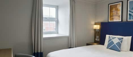 A hotel room with a bed, a desk, a chair, a nightstand, a lamp, and two framed pictures on the wall.