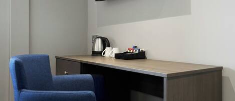 A hotel room with a desk, a chair, a kettle, a phone, and a small black tray with items.