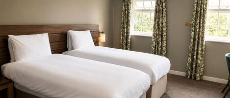 A hotel room with two beds, a wooden headboard, a nightstand, a desk, and a window with curtains.