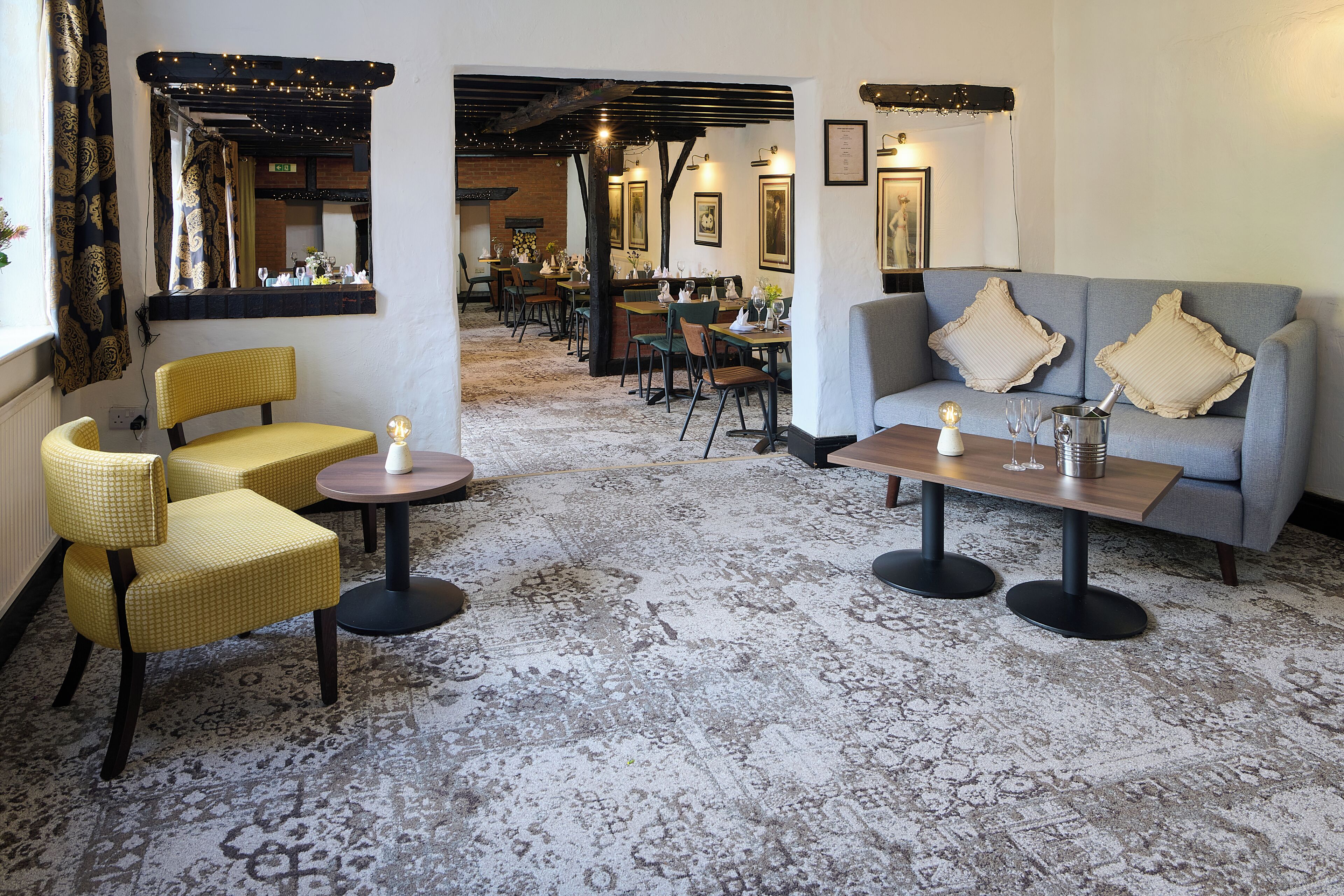 Un hall de hotel con un área de descanso que incluye sillas amarillas, un sofá gris y una mesa de madera. Hay cuadros enmarcados en las paredes y una pared de ladrillo al fondo.
