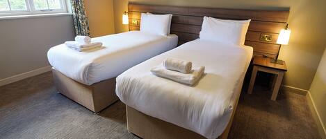 A hotel room with two single beds, each with a rolled towel, a wooden headboard, bedside lamps, and a carpeted floor.