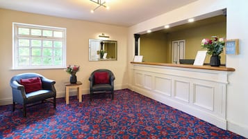 Zona de recepción de un hotel con alfombra estampada, dos sillones, una mesita con un jarrón de flores, un espejo y un mostrador de recepción con un cartel.