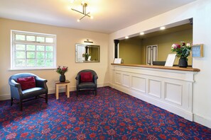 A hotel reception area with a patterned carpet, two armchairs, a small table with a vase of flowers, a mirror, and a reception desk with a sign. - Marston Farm Hotel (Sutton Coldfield)