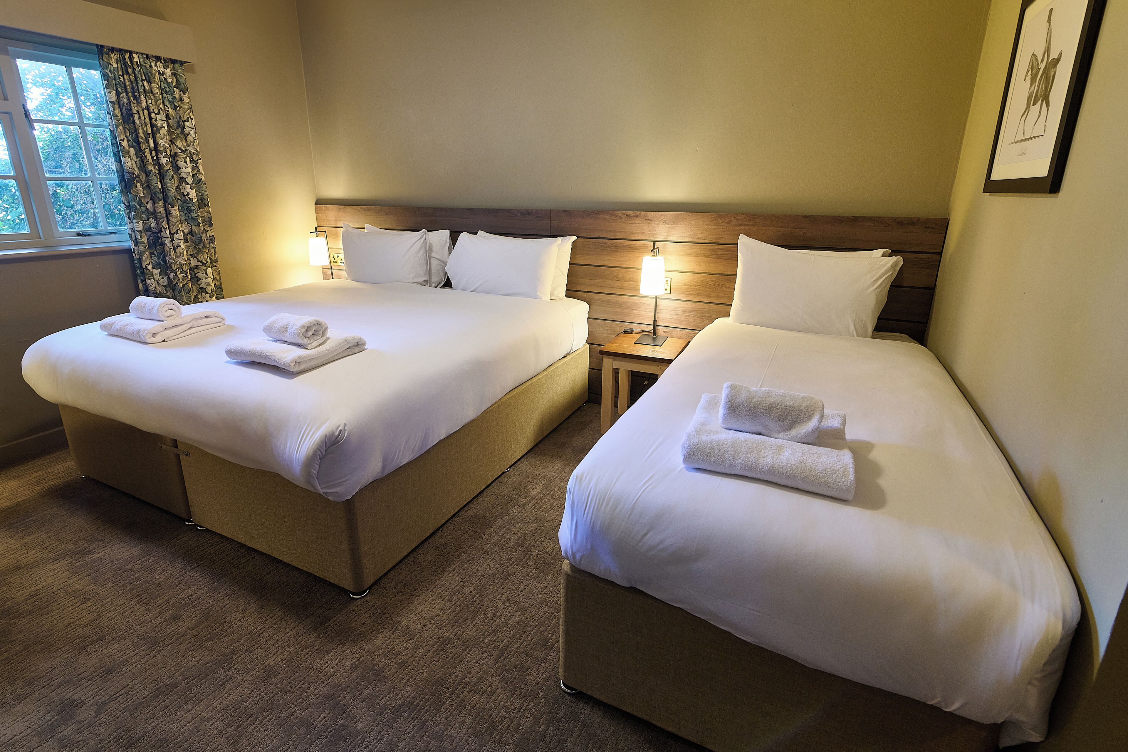Une chambre d’hôtel avec deux lits, chacun équipé de draps et de serviettes blancs, d’une tête de lit en bois, d’une table de chevet avec une lampe et d’une fenêtre avec des rideaux.