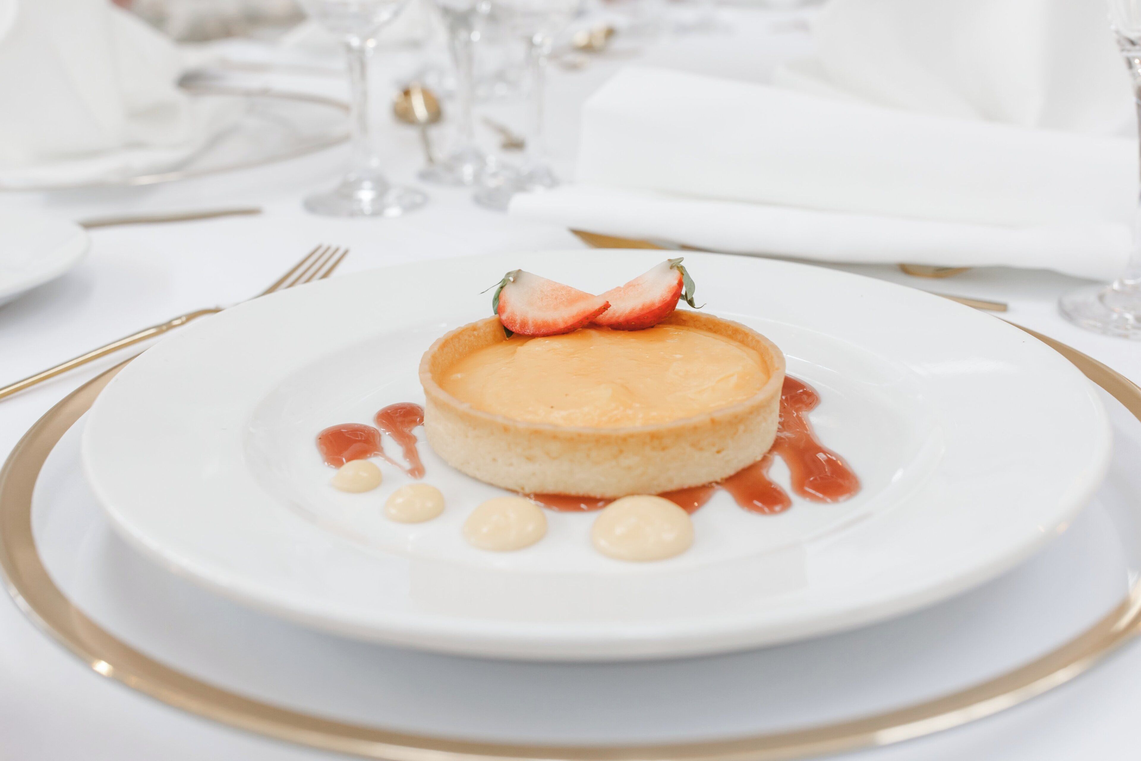 Dessert auf einem weißen Teller mit goldener Kante, verziert mit Erdbeeren und Soße, auf einem Tisch mit weißem Geschirr und Gläsern.