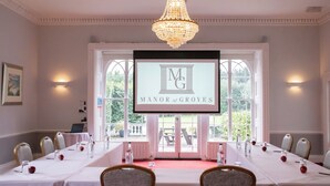 A conference room with a long table, chairs, a chandelier, and a screen displaying 'MANOR of GROVES'. - Manor of Groves Hotel, Golf & Health Club (Sawbridgeworth)