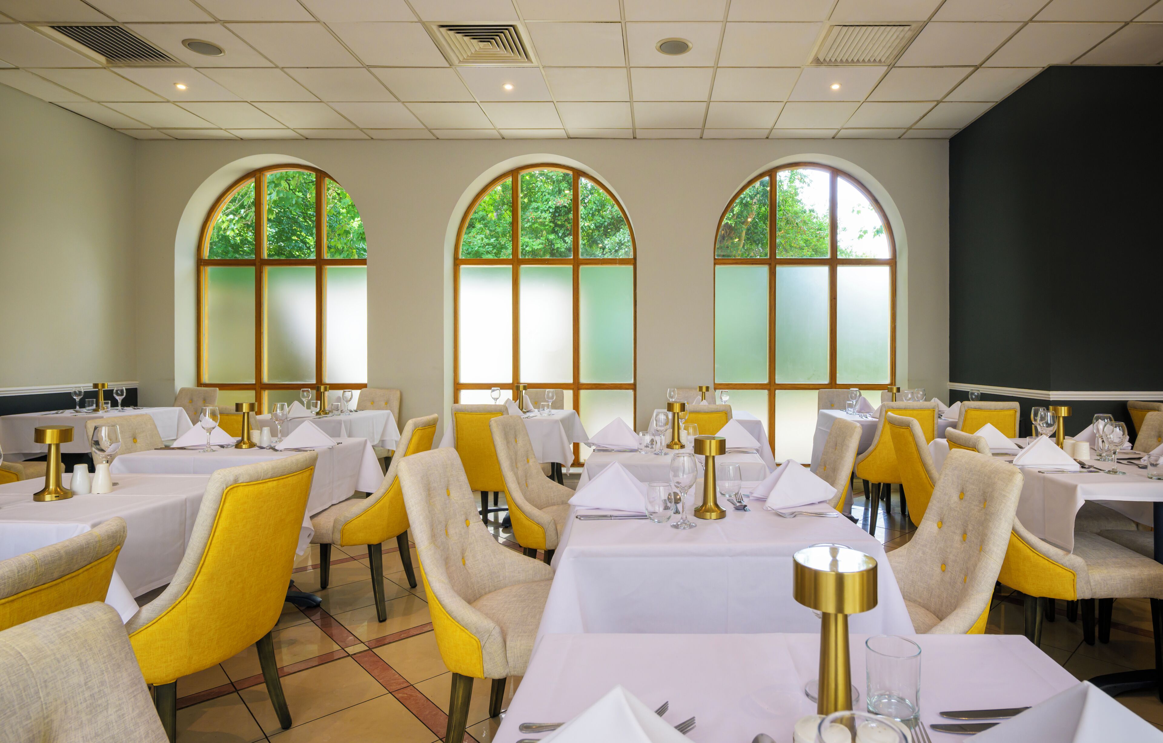 A dining area with tables set for service, chairs upholstered in yellow, and large arched windows with frosted glass.