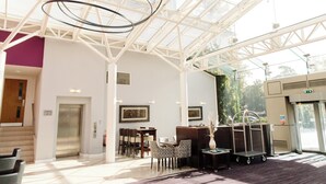 A spacious lobby with a high ceiling, a reception desk, seating area, and a glass elevator. - Manor of Groves Hotel, Golf & Health Club (Sawbridgeworth)