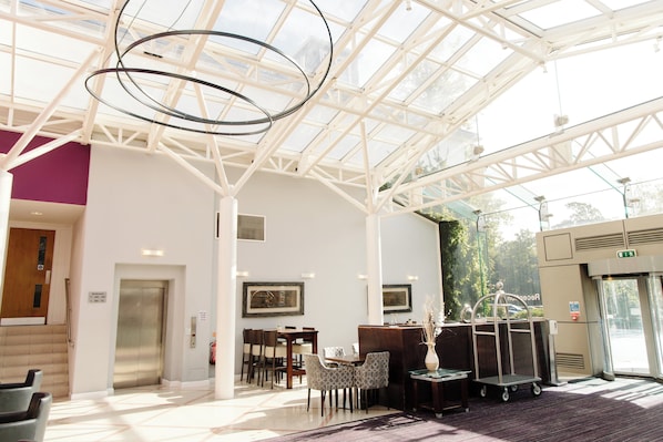 A spacious lobby with a high ceiling, a reception desk, seating area, and a glass elevator.