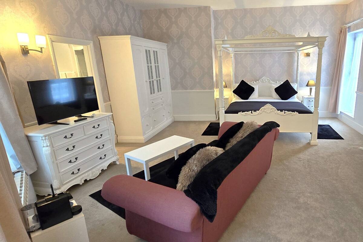 A bedroom with a four-poster bed, a television, a wardrobe, a sofa, and a desk with a chair.