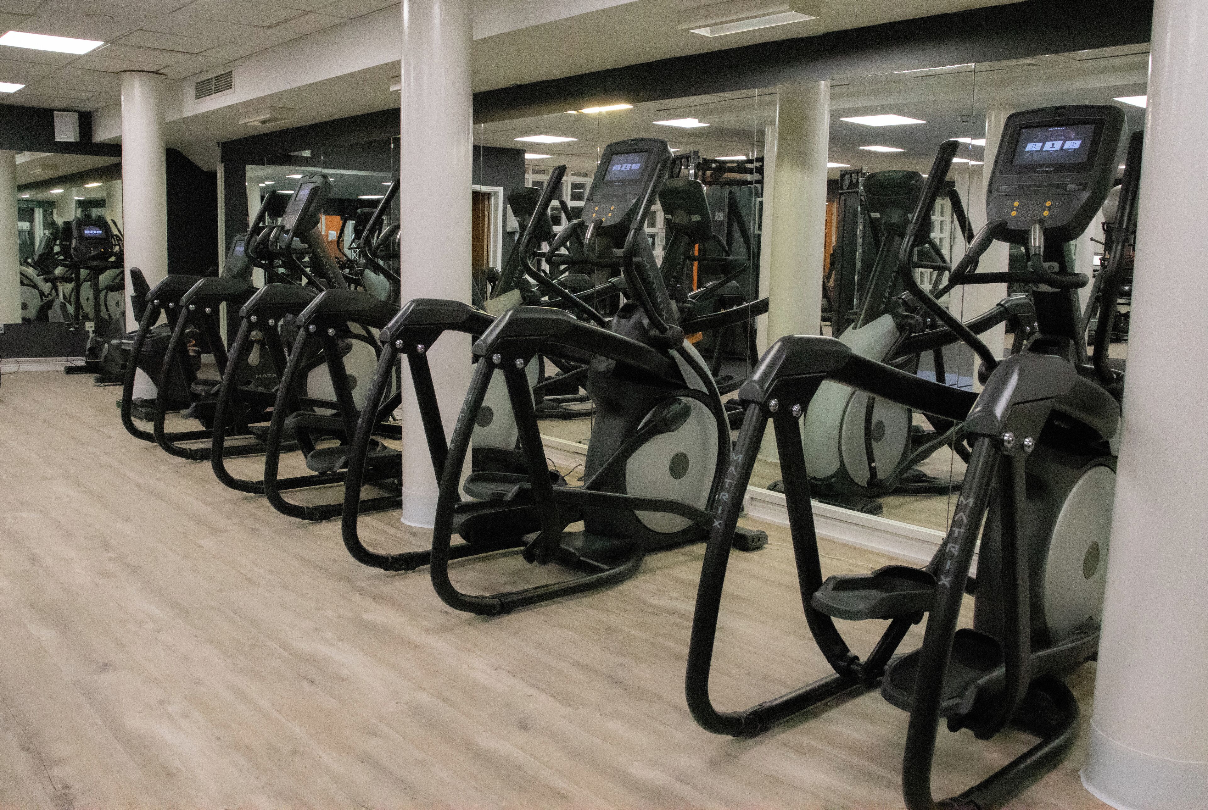 En rad med motionscyklar i ett gym med trägolv och vita pelare.
