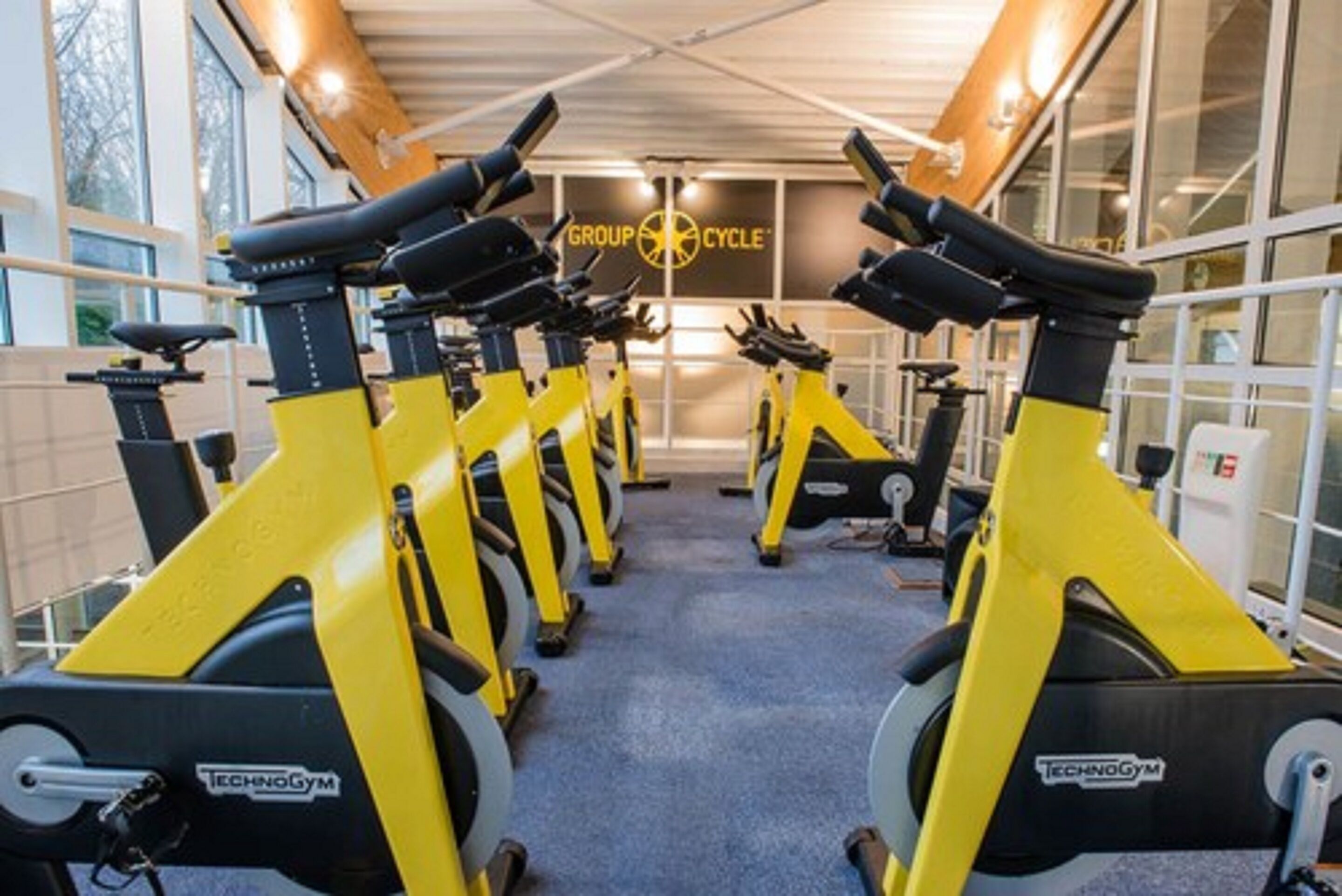 A gym with stationary bikes arranged in rows, a sign reading "GROUP CYCLE", and a logo "TECHNOGYM".