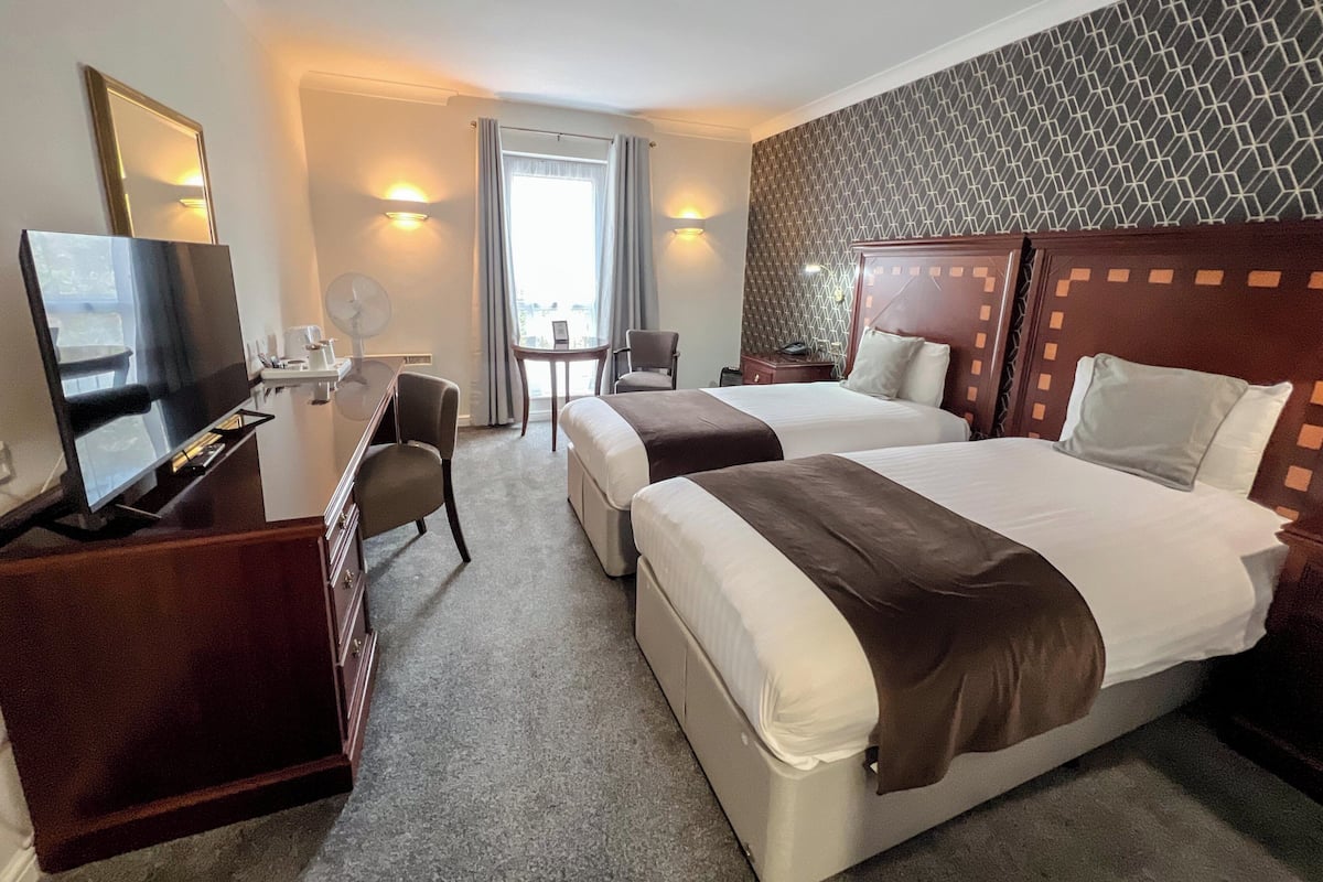A hotel room with two beds, a desk with a TV, a chair, a small table, and a window with curtains.