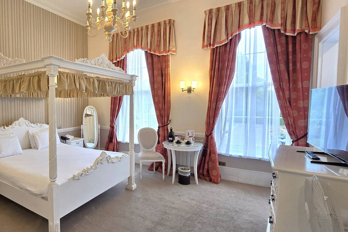 A four-poster bed with a canopy, a bedside table, a chair, a small table with a lamp, and a window with curtains.