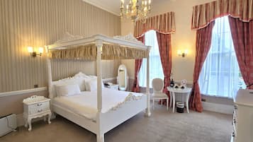 A four-poster bed with a canopy, a bedside table, a chair, a small table with a lamp, and a window with curtains.