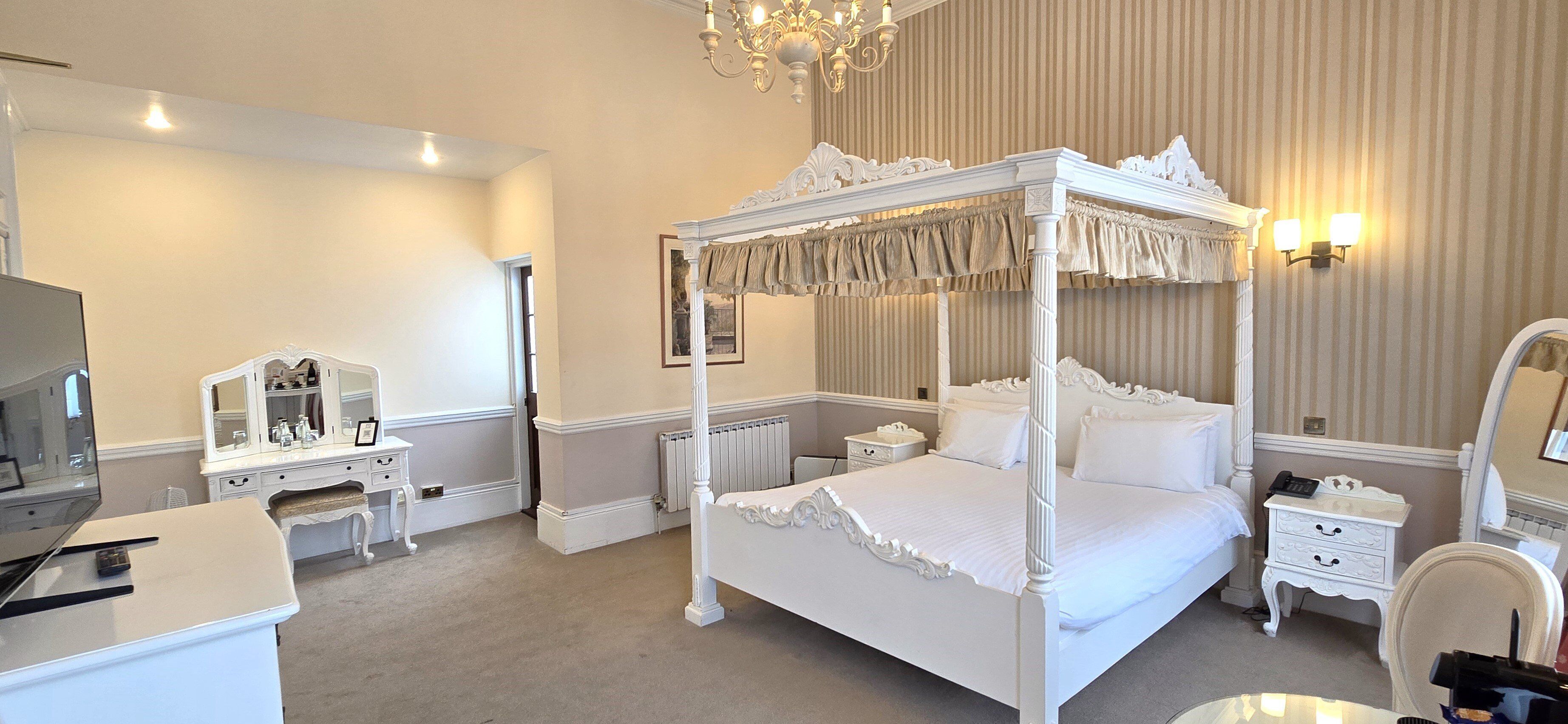 A four-poster bed with a canopy, a vanity with a mirror, a bedside table, a chair, and a television.