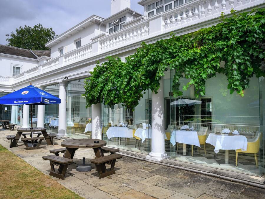 An outdoor seating area with tables and benches, a blue umbrella, and a building with white columns and a vine-covered wall.