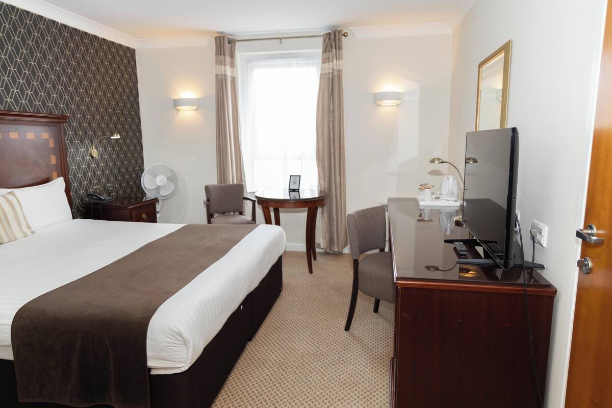A hotel room with a large bed, a desk with a television, a chair, a small table, and a window with curtains.
