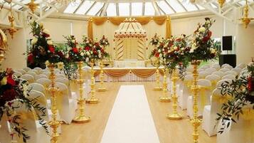 Un espacio interior con un pasillo central, sillas blancas a cada lado y elementos decorativos dorados.