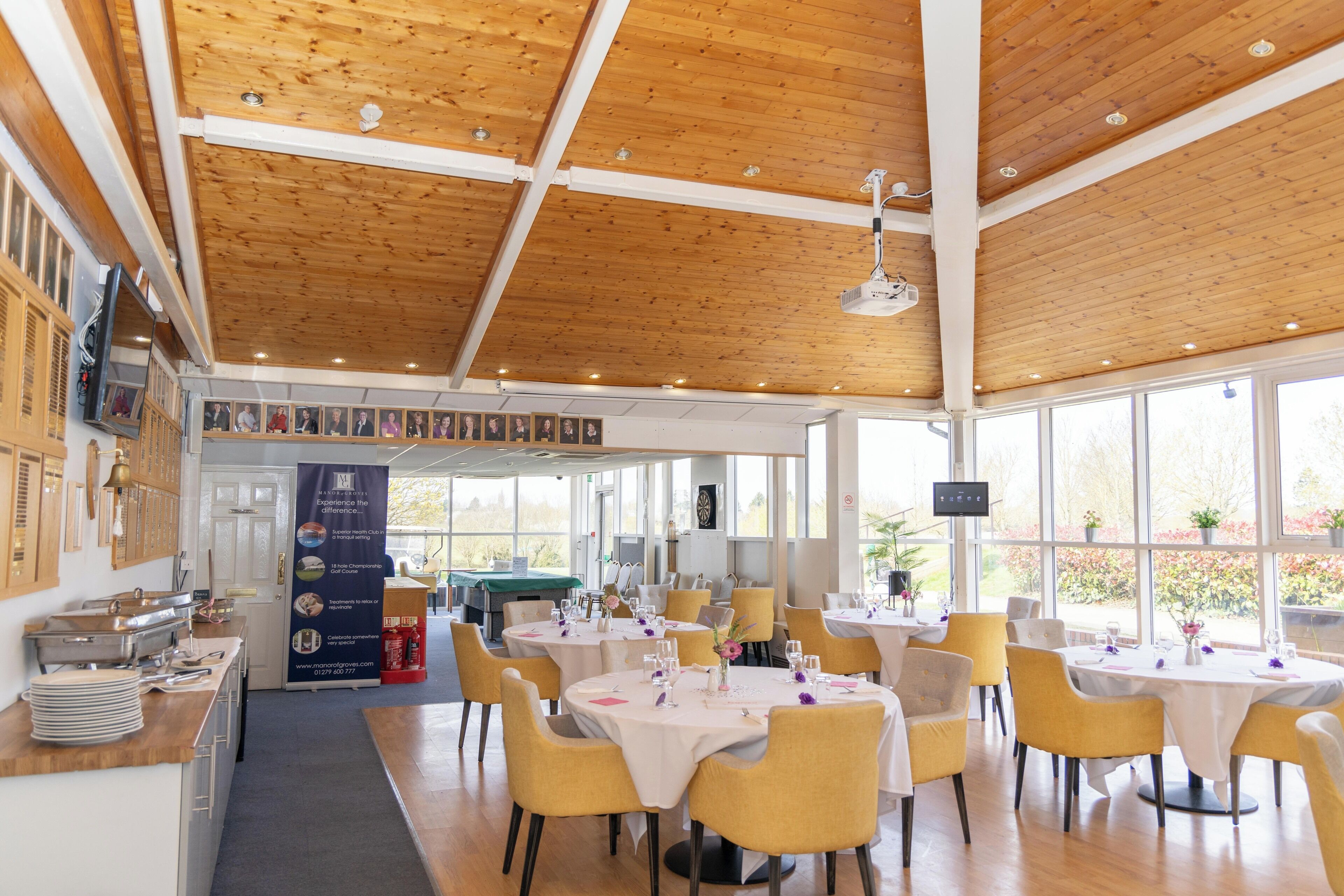 A restaurant with wooden ceilings and large windows. Tables are set with white tablecloths and yellow chairs.