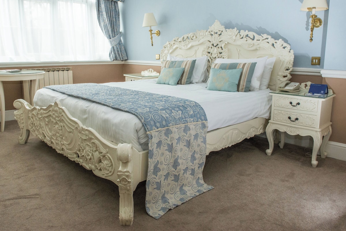 A classic four-poster bed with a blue and white patterned bedspread, a white nightstand with a lamp, and a small table with a book and a phone.