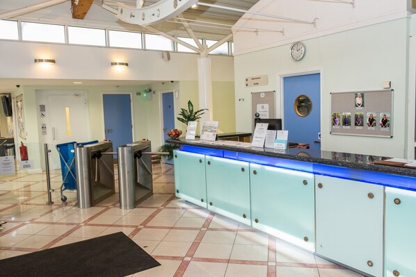 Zona de recepción con puerta azul, mostrador de recepción, tablón de anuncios y un reloj en la pared.