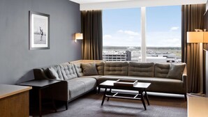 A modern hotel room with a large sectional sofa, a small table, a desk, and a large window offering a view of the airport.