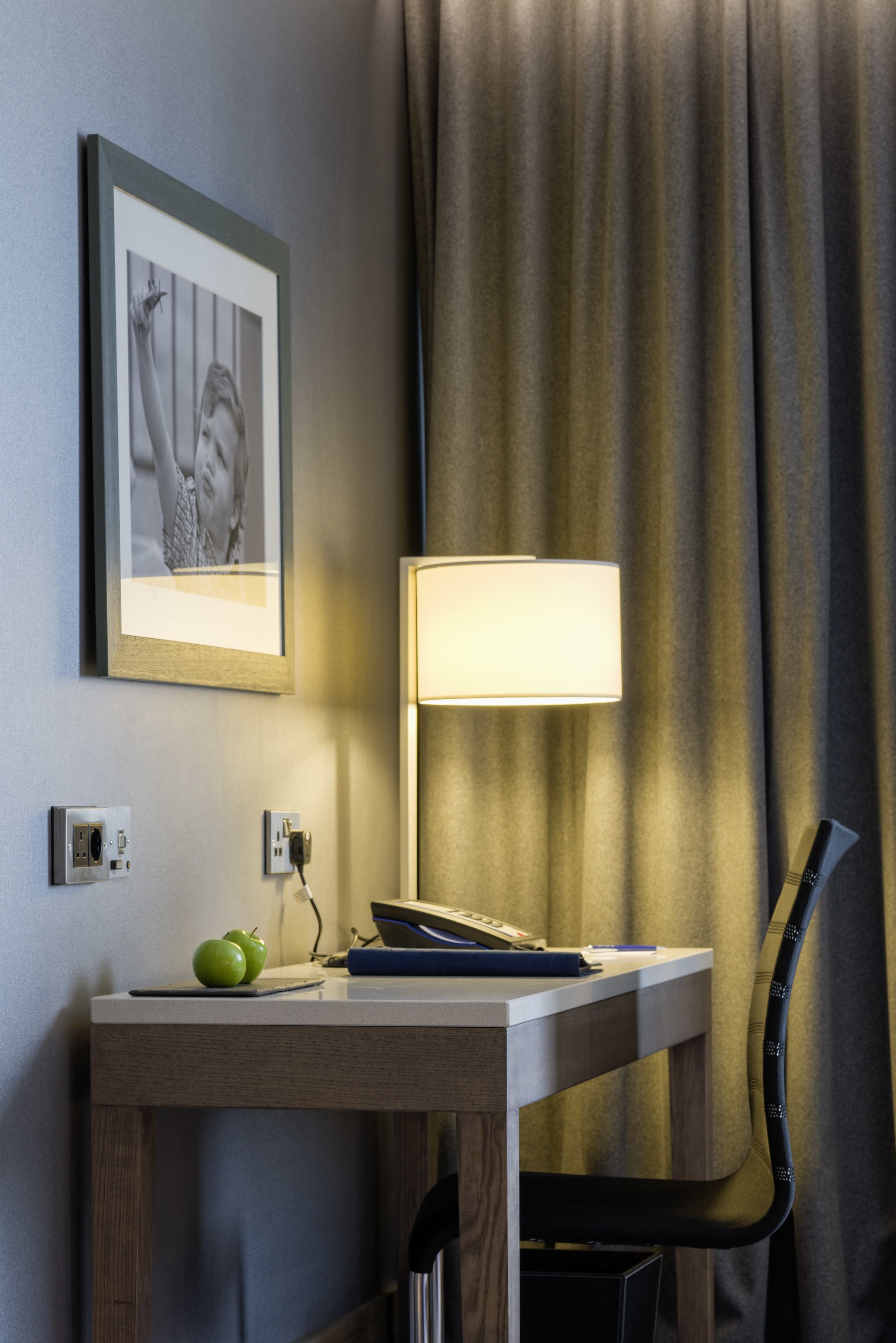 Quarto de hotel com escrivaninha, luminária, quadro na parede e cortina.