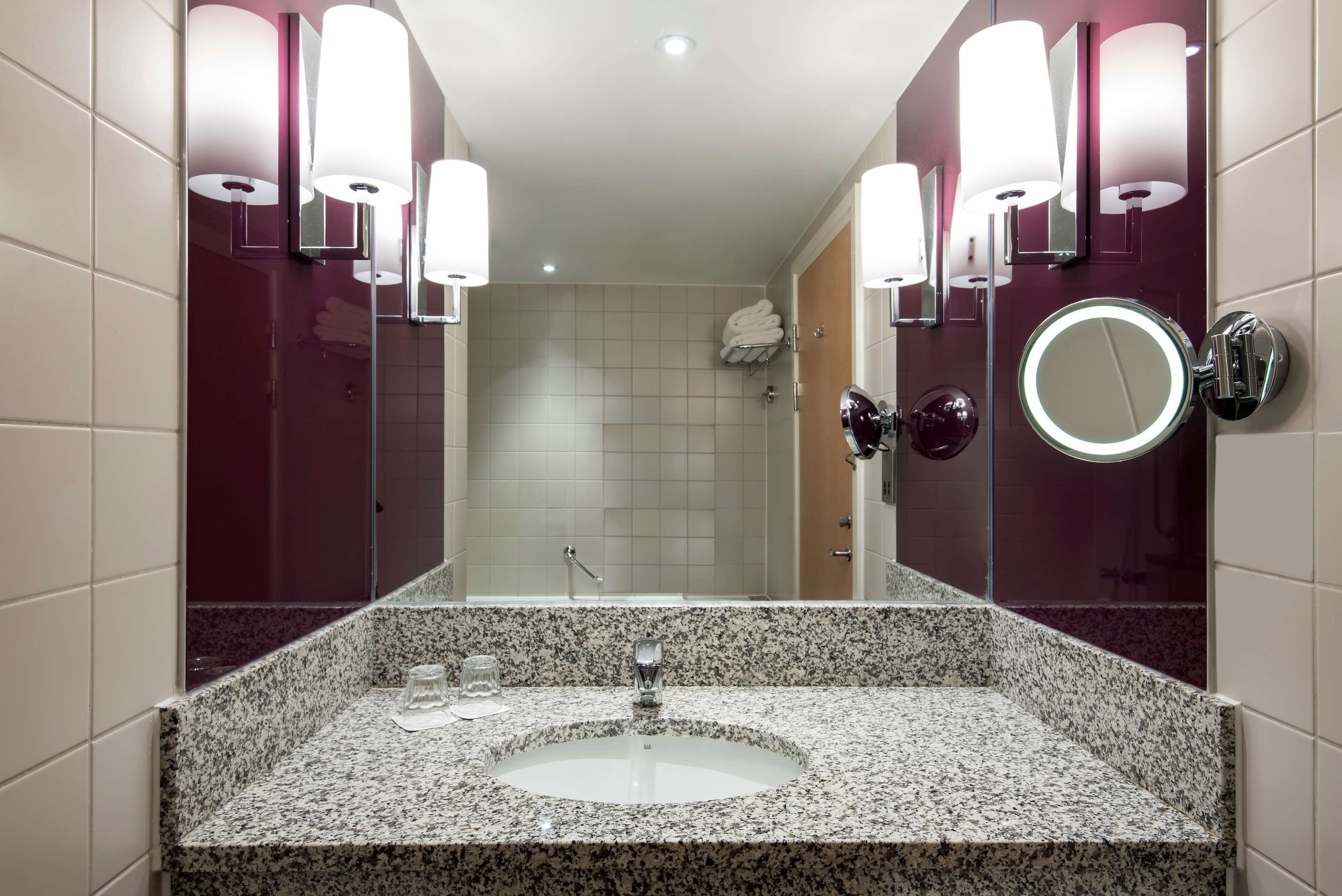 A bathroom with a granite countertop sink, a mirror with a round mirror above it, and wall-mounted lights.