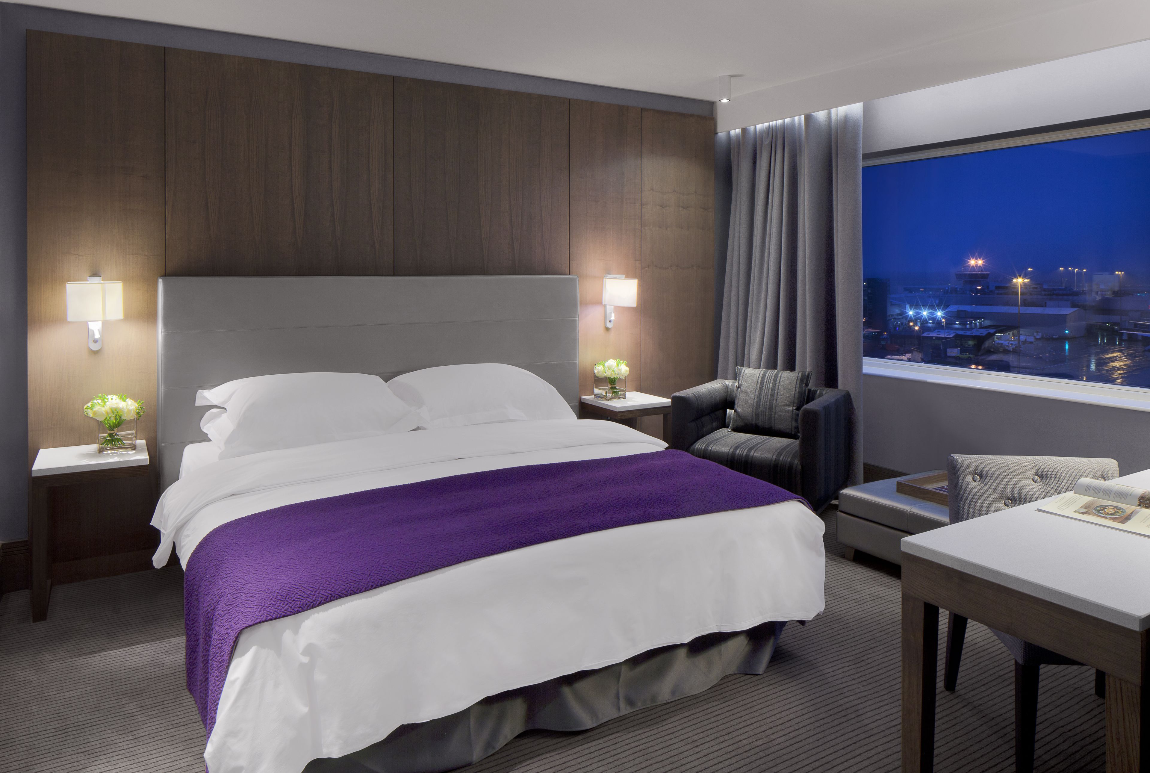 Una camera d'albergo con un letto grande, una scrivania, una sedia, un comodino con una lampada e vista sulla città illuminata.