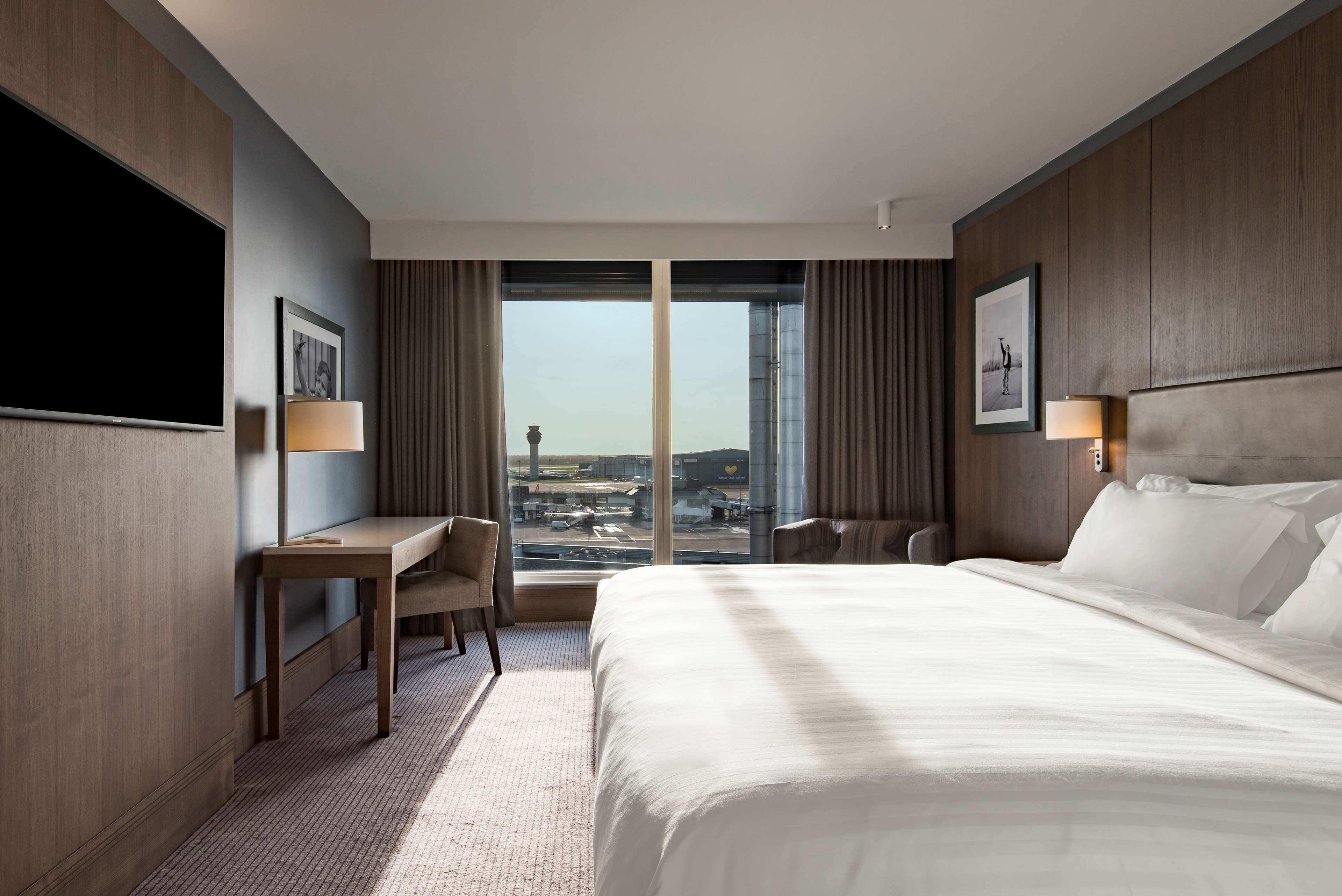 A hotel room with a large bed, a desk, a chair, a TV, and a view of the city through large windows.